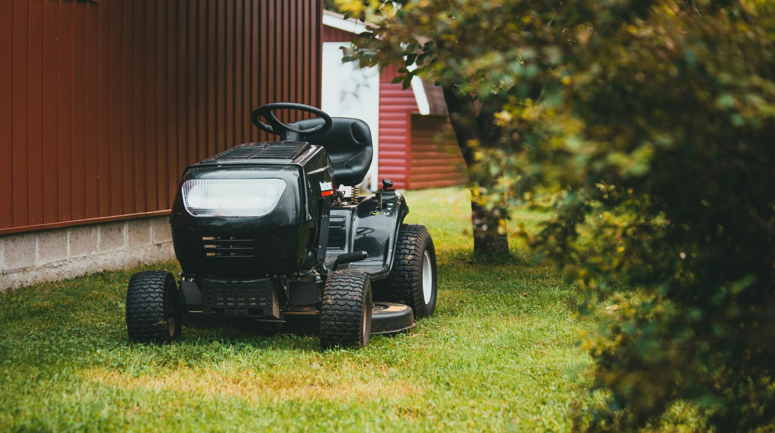 A black ride-on lawnmower parked on green grass near a red barn during summer.